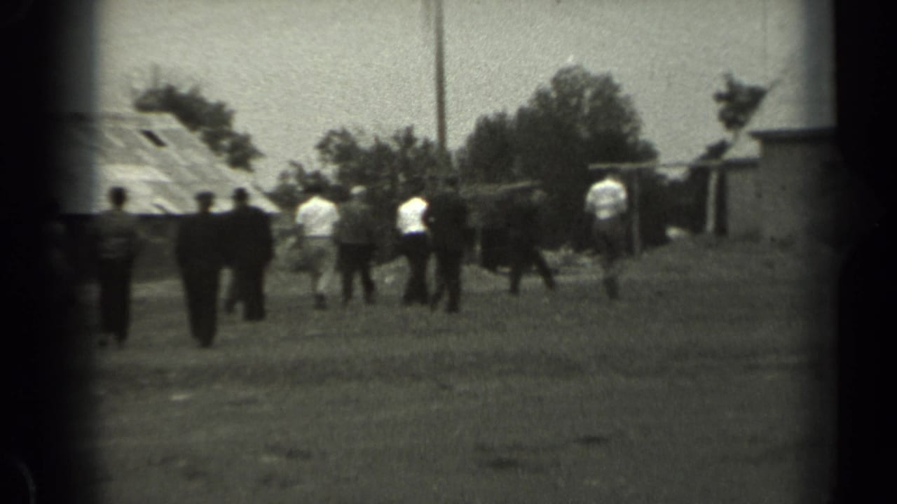1961 Missouri home-movie frame showing a group of people walking across a grassy field near barns and a utility pole.