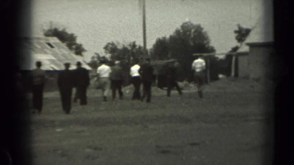 1961 Missouri home-movie frame showing a group of people walking across a grassy field near barns and a utility pole.