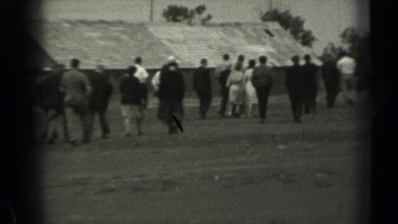 Black-and-white 8mm frame from 1961 Missouri showing people walking away across open farm ground near barn roofs and trees.