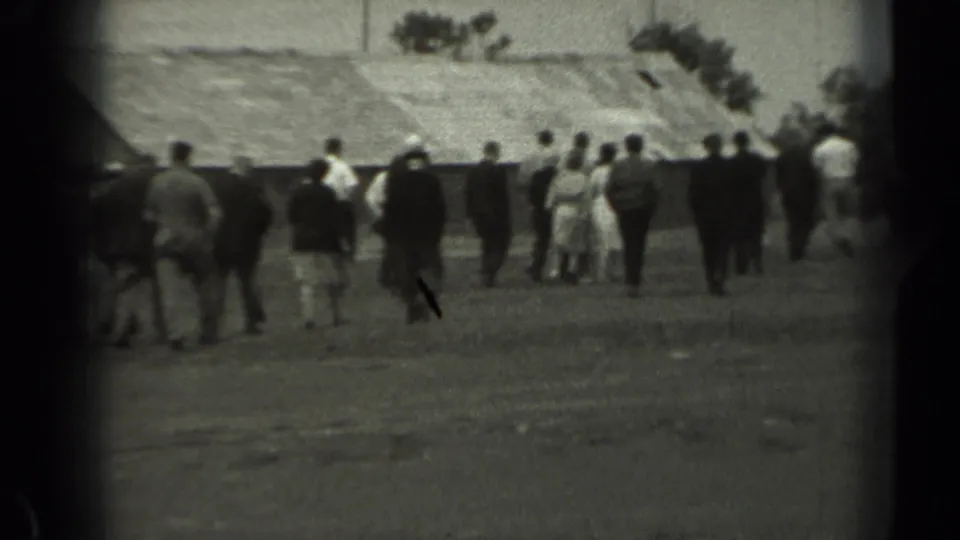 Black-and-white 8mm frame from 1961 Missouri showing people walking away across open farm ground near barn roofs and trees.