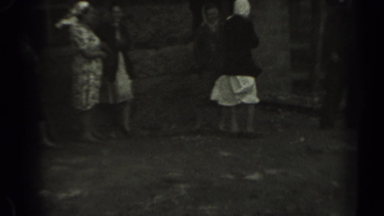 In 1961 Missouri home-movie footage, adults gather outdoors near grass, trees, and a wooden fence as a woman in a patterned dress stands at left.