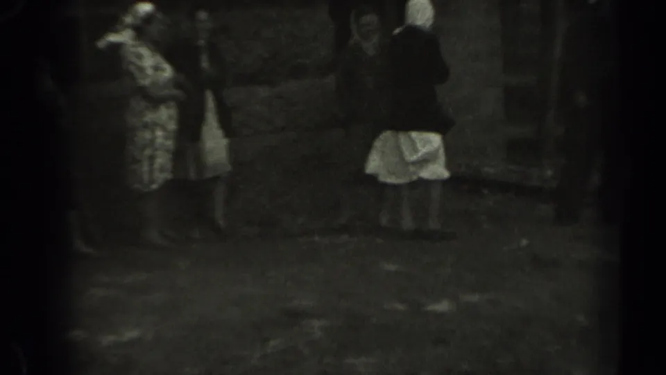 In 1961 Missouri home-movie footage, adults gather outdoors near grass, trees, and a wooden fence as a woman in a patterned dress stands at left.