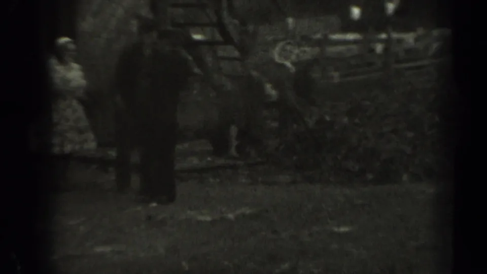 A woman in a patterned dress and a man in dark clothing stand near brush or leaves by a wooden fence in rural Missouri footage from 1961.
