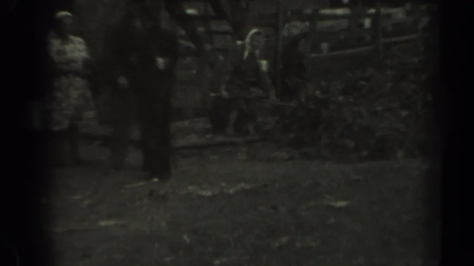 Adults appear around a pile of leaves or brush, with a wooden fence and steps behind them in this grainy 1961 Missouri home-movie frame.