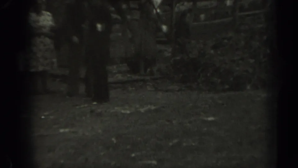 The frame shows a woman in a patterned dress and a man in dark clothing near a fence and ground leaves in rural Missouri, 1961.