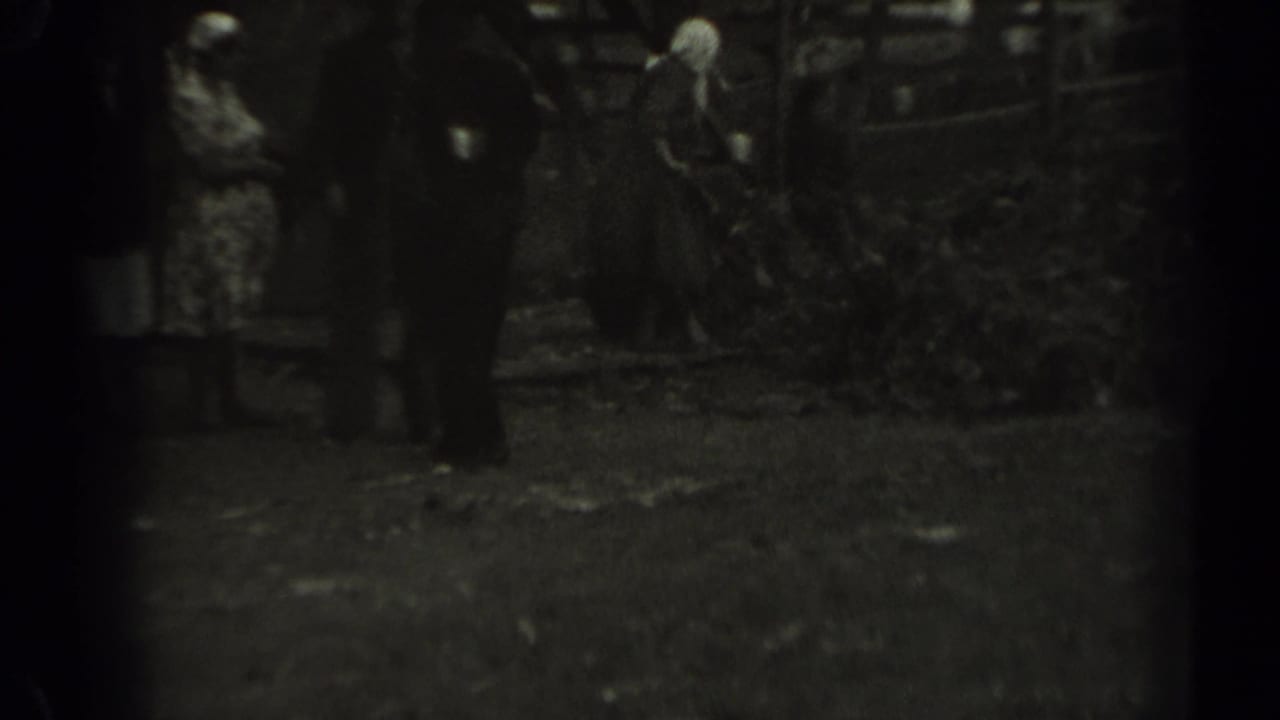 Grainy 1961 Missouri home-movie frame with adults gathered as a woman in a headscarf works with a rake near leaves.