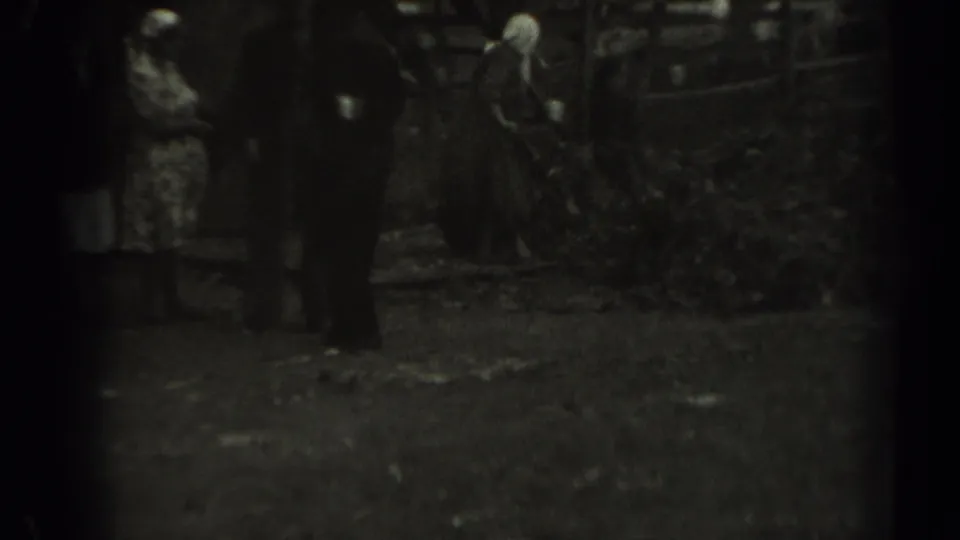 Grainy 1961 Missouri home-movie frame with adults gathered as a woman in a headscarf works with a rake near leaves.