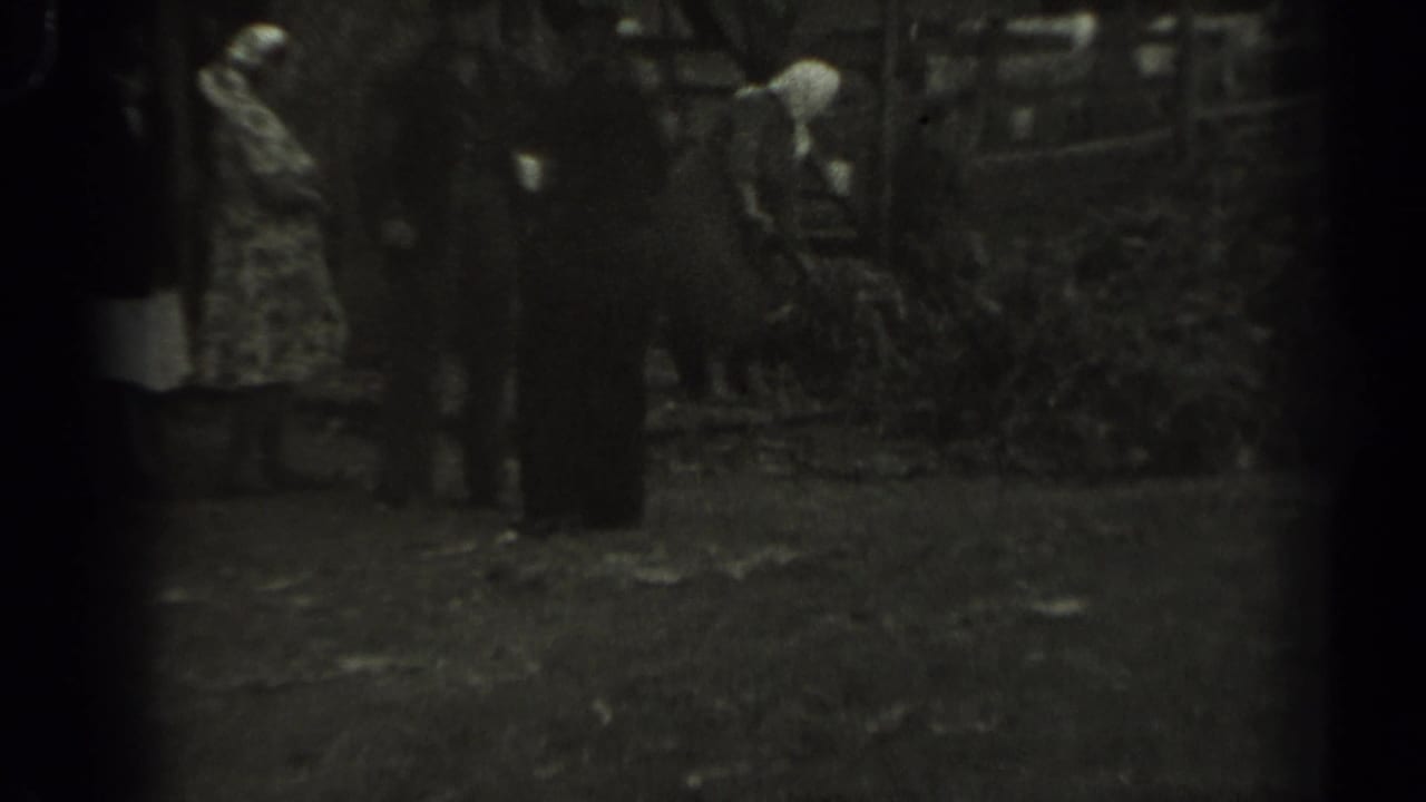 1961 Missouri backyard footage showing a woman in a white headscarf raking leaves beside standing adults and a rustic fence.