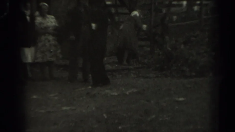 1961 Missouri 8mm frame showing adults in a rural yard beside a wooden animal pen, including a woman in a patterned dress.
