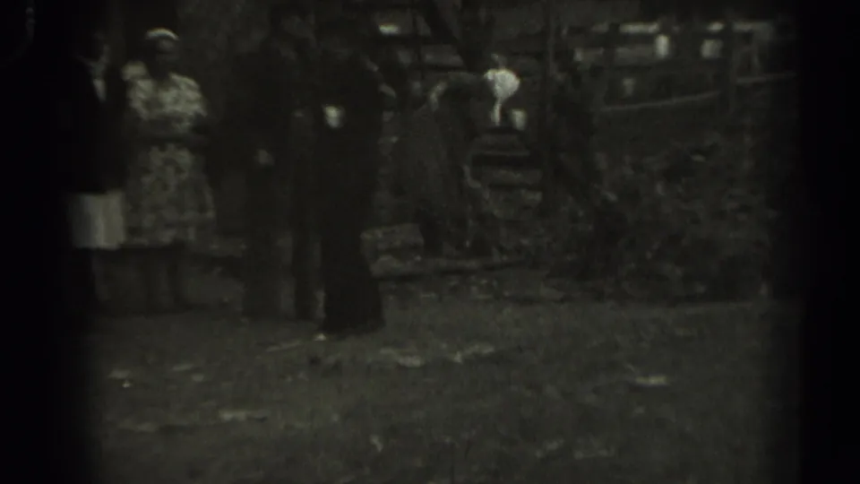 Grainy 1961 Missouri home-movie frame with adults near a wooden pen and a person in a light head covering moving in the background.