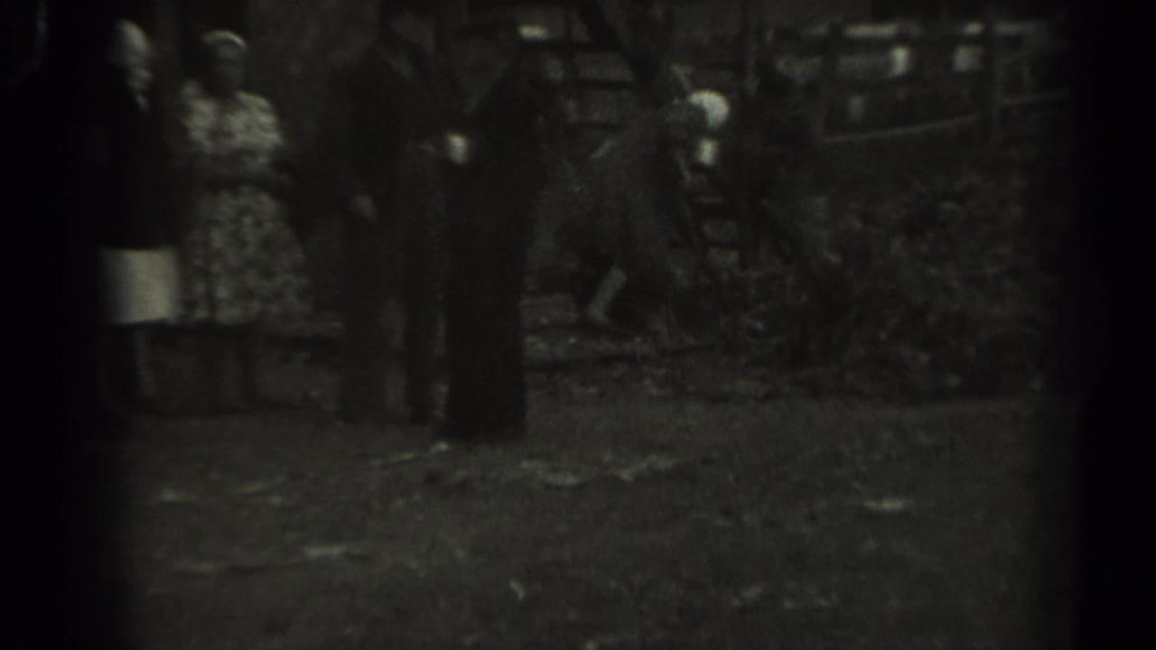 1961 Missouri 8mm frame with a patterned-dress woman at left and several adults clustered near a wooden animal pen.