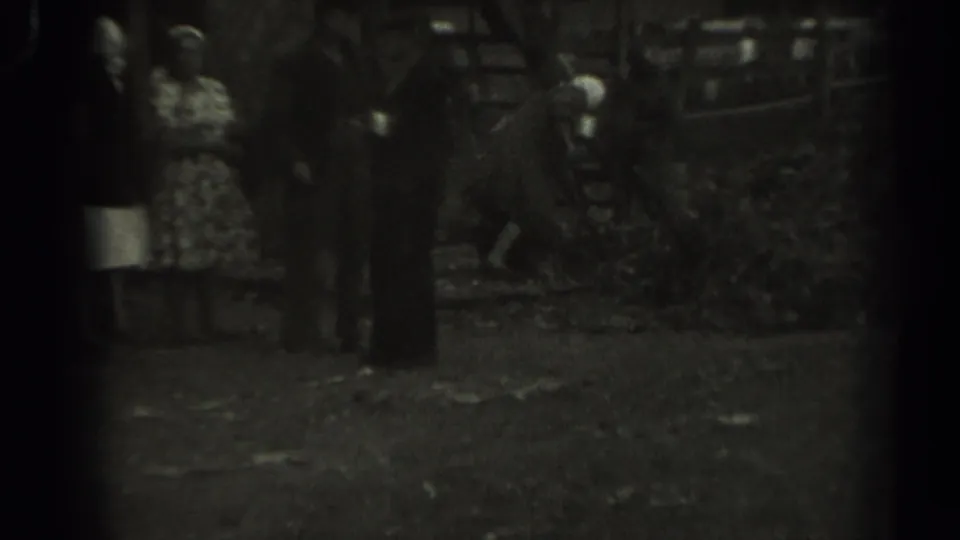 1961 Missouri 8mm frame with a patterned-dress woman at left and several adults clustered near a wooden animal pen.
