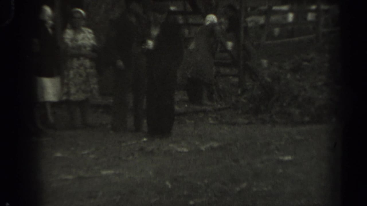 Soft black-and-white 1961 Missouri frame showing adults near a wooden animal pen, with foliage and yard ground visible around them.