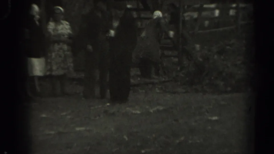 Soft black-and-white 1961 Missouri frame showing adults near a wooden animal pen, with foliage and yard ground visible around them.