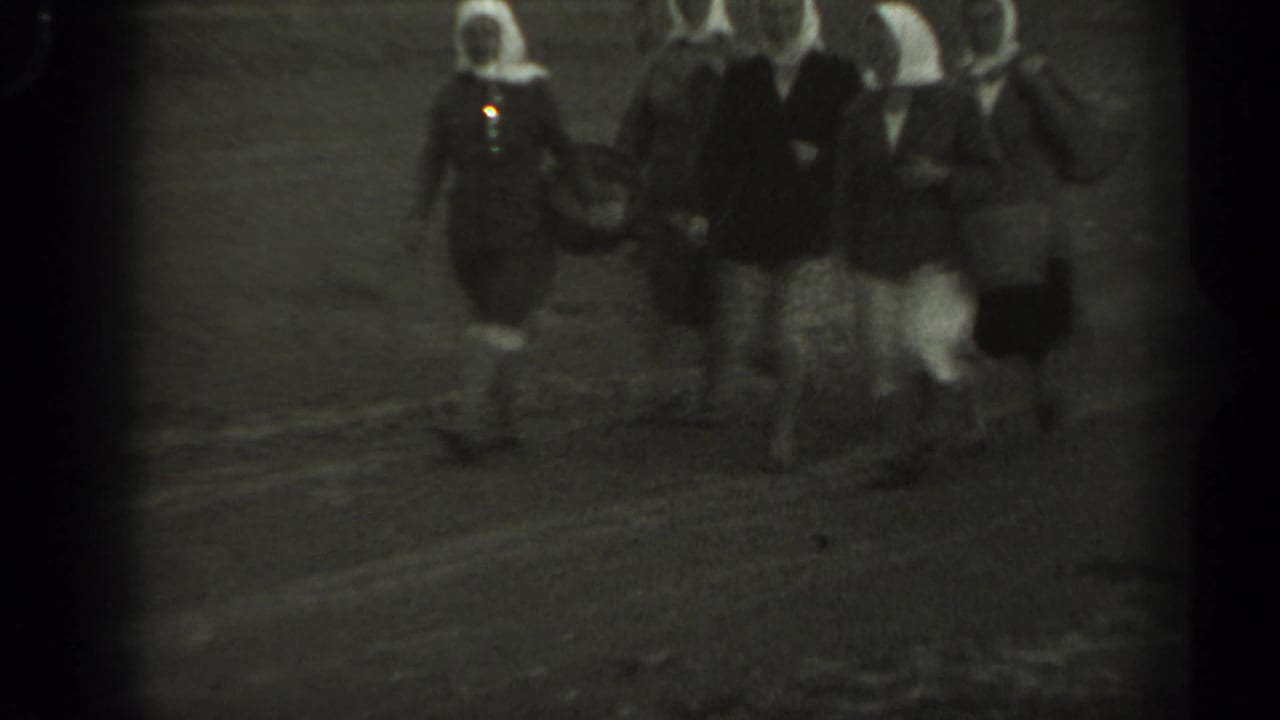 Passengers walk across a tarmac or open airport area in soft black-and-white footage.