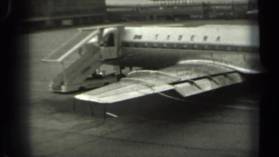 Black-and-white 8mm frame of a Sabena passenger plane at a Missouri airport, showing boarding stairs and the aircraft wing in the foreground.