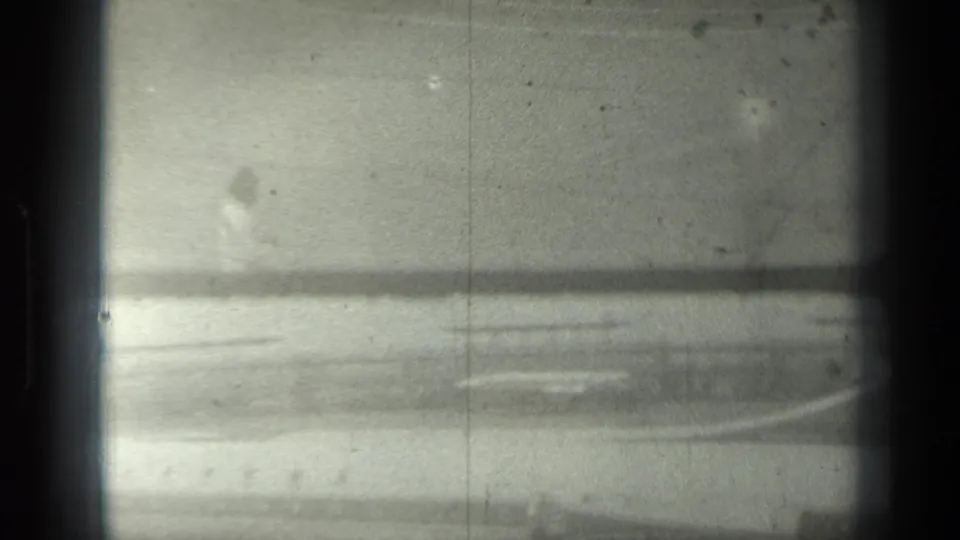 Soft, low-contrast 8mm frame showing blurred airport pavement or ramp surface with window edge and film wear.