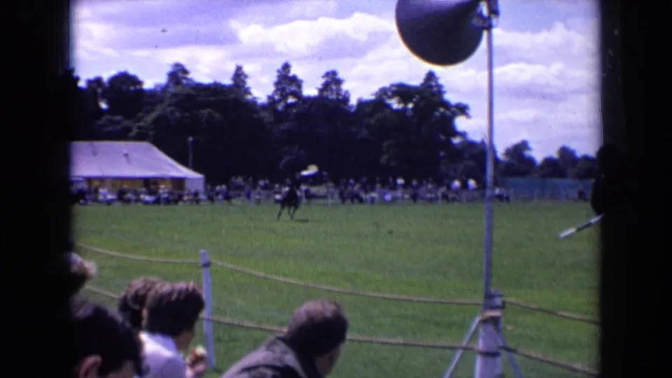 1963 England home-movie frame of a black horse and rider on a grass field, with a crowd, white tent, trees, and rope barrier visible.