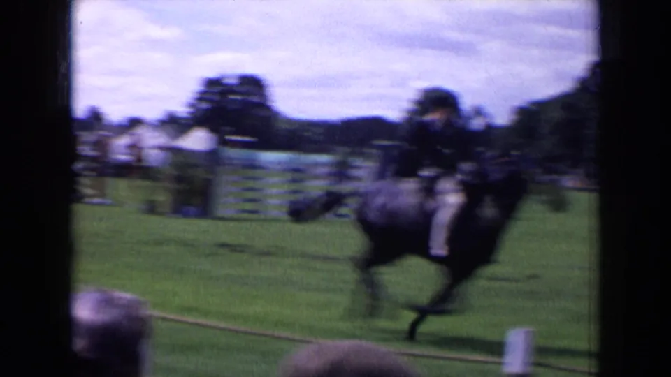 Motion-blurred 8mm frame of a dark horse and rider passing close to the camera at an English park equestrian event.