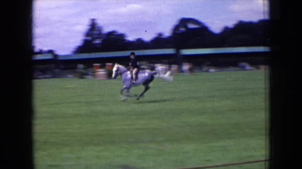 White horse and rider gallop across a grassy English park field in 1963, with trees and a long low structure behind them.
