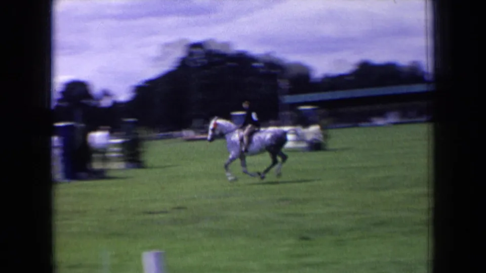 In England 1963, a rider in dark attire gallops a dapple gray horse across a grass field with fencing and tents beyond.