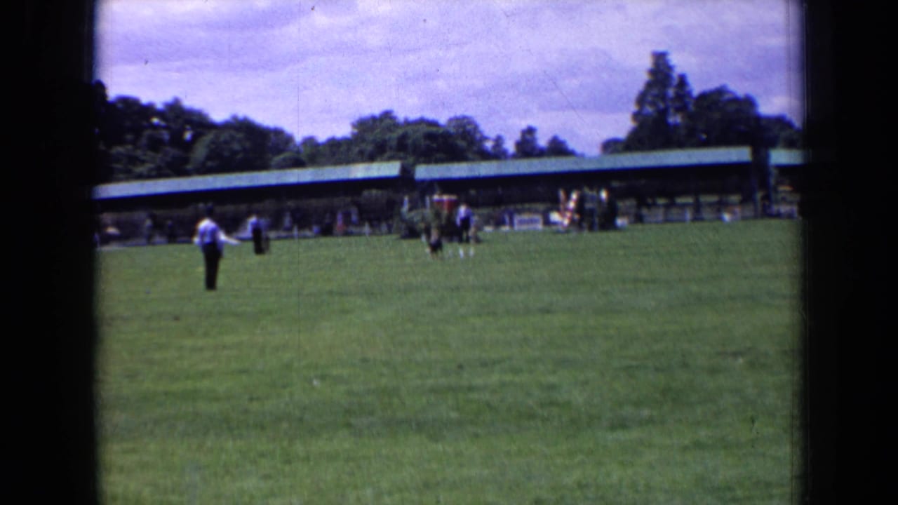 In England 1963 home-movie footage, a man in a light shirt stands on grass near distant spectators and a green-roofed pavilion.