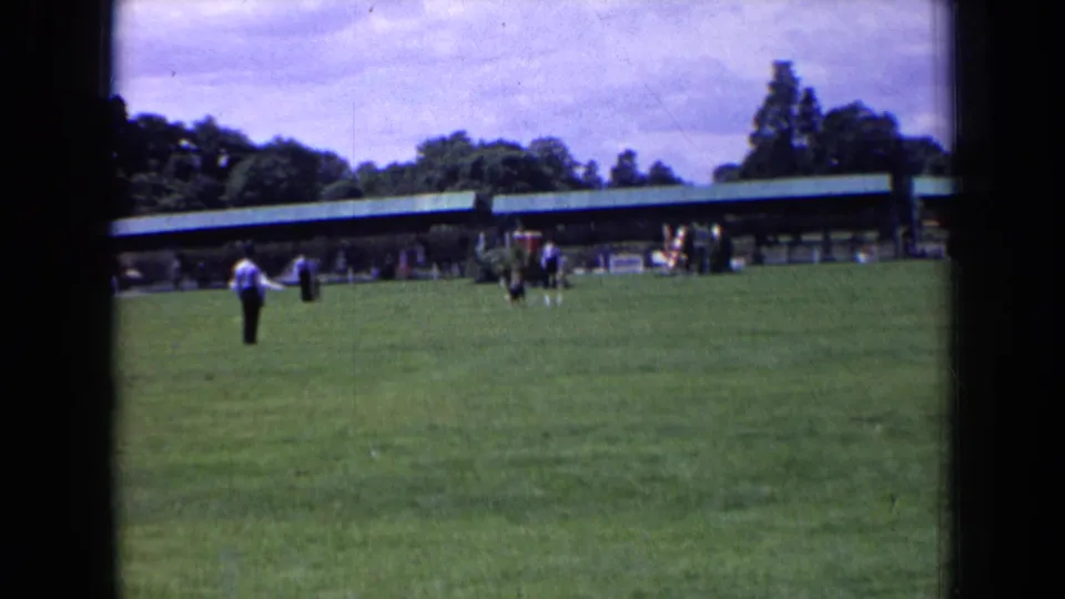 In England 1963 home-movie footage, a man in a light shirt stands on grass near distant spectators and a green-roofed pavilion.