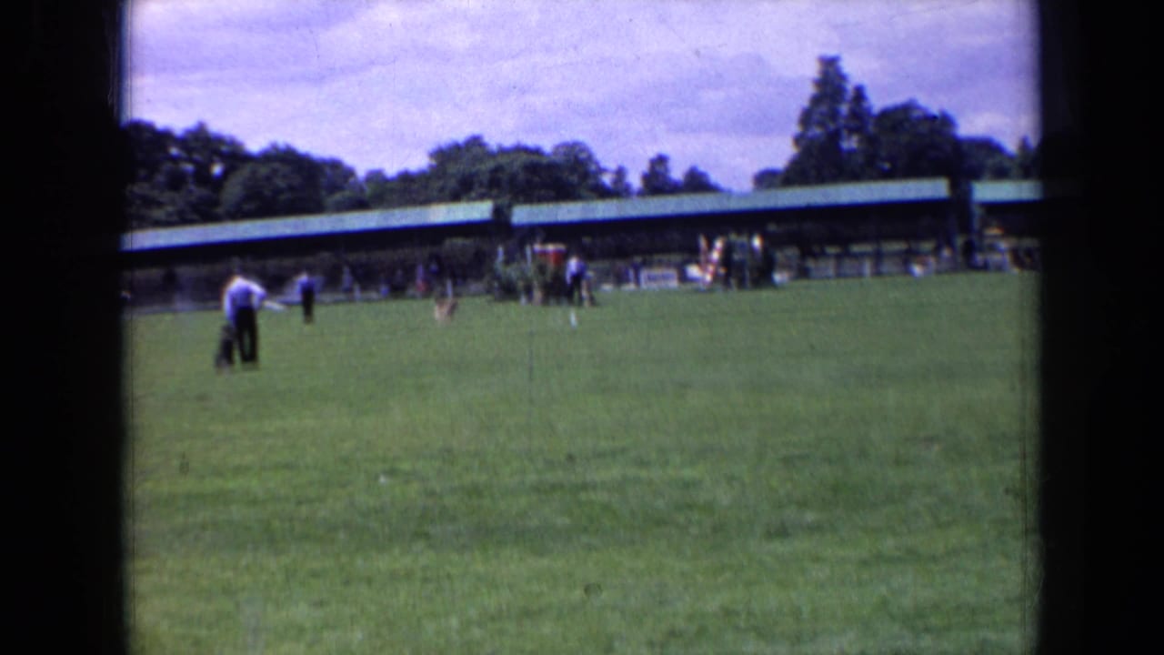 1963 England 8mm frame showing a dog crossing a grass field with spectators and a long pavilion in the background.