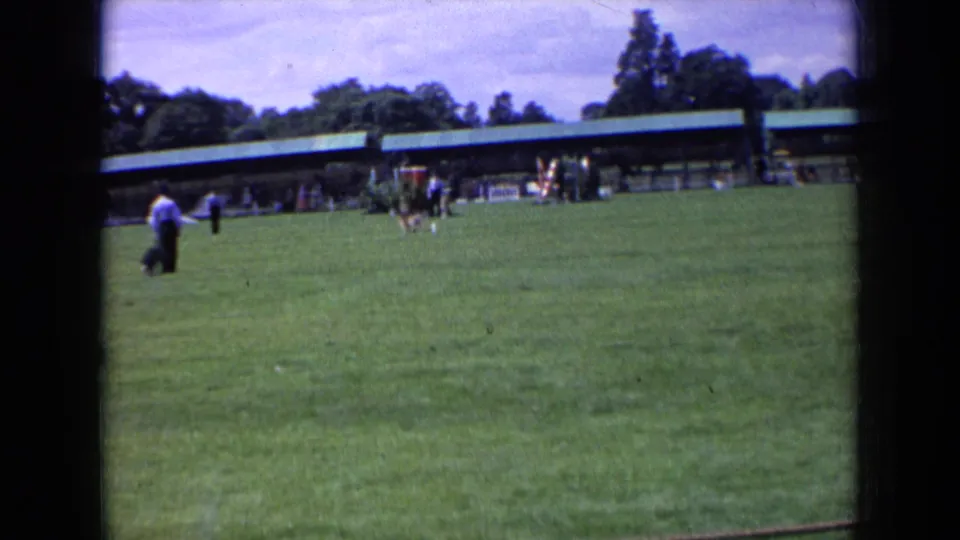 Soft 8mm frame from 1963 England showing a dog on a lawn with people watching near a long covered structure.