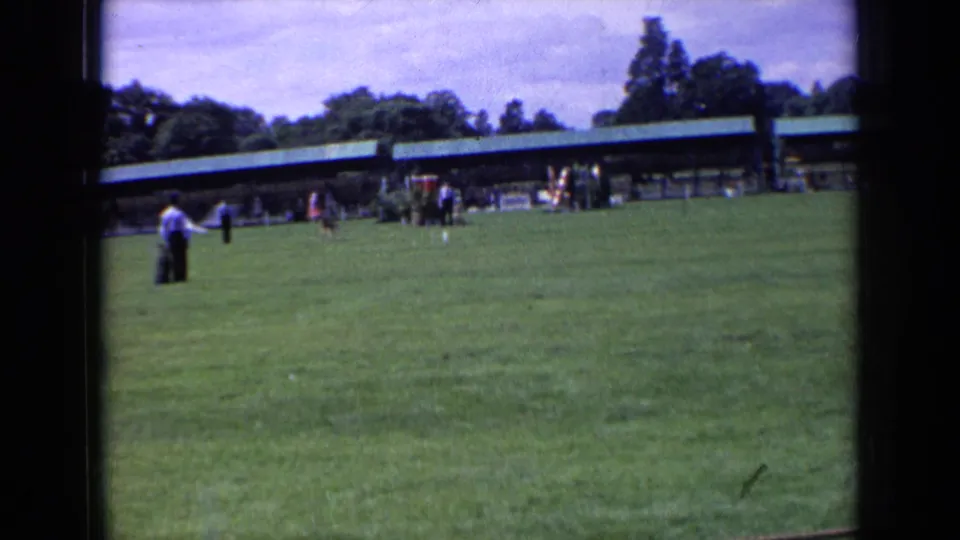 1963 England 8mm frame of a wide field scene with a small dog, distant spectators, trees, and a long pavilion.