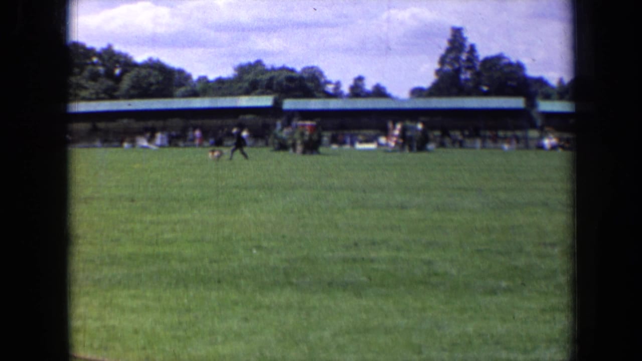 1963 England park scene with a dark-clothed runner and black dog moving across the field before a green-roof pavilion.