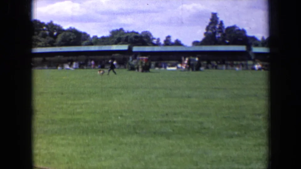 1963 England park scene with a dark-clothed runner and black dog moving across the field before a green-roof pavilion.