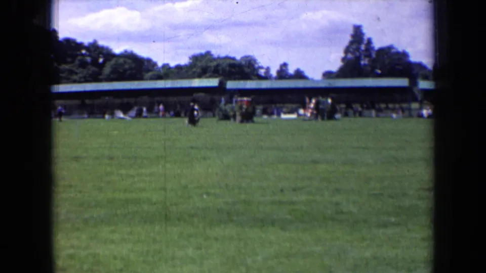 Soft 8mm frame of a 1963 England park, with the runner and black dog set against a long pavilion and tree-lined background.