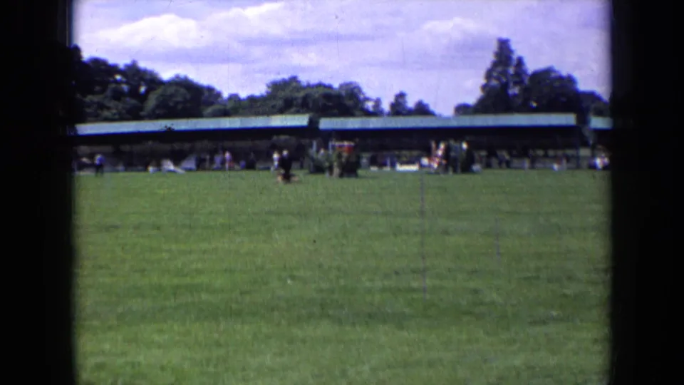 1963 England home-movie frame showing a runner and black dog in a wide public park field with a green-roof pavilion behind.