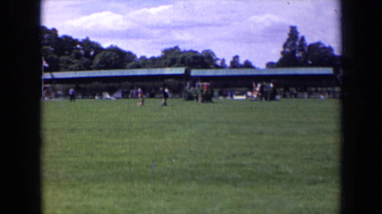 Wide grass field with distant people near a long pavilion under cloudy sky.