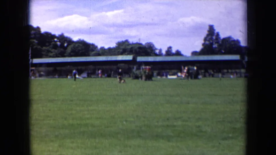 1963 England 8mm frame with a green field in the foreground, small distant figures near a long pavilion, and trees on the horizon.
