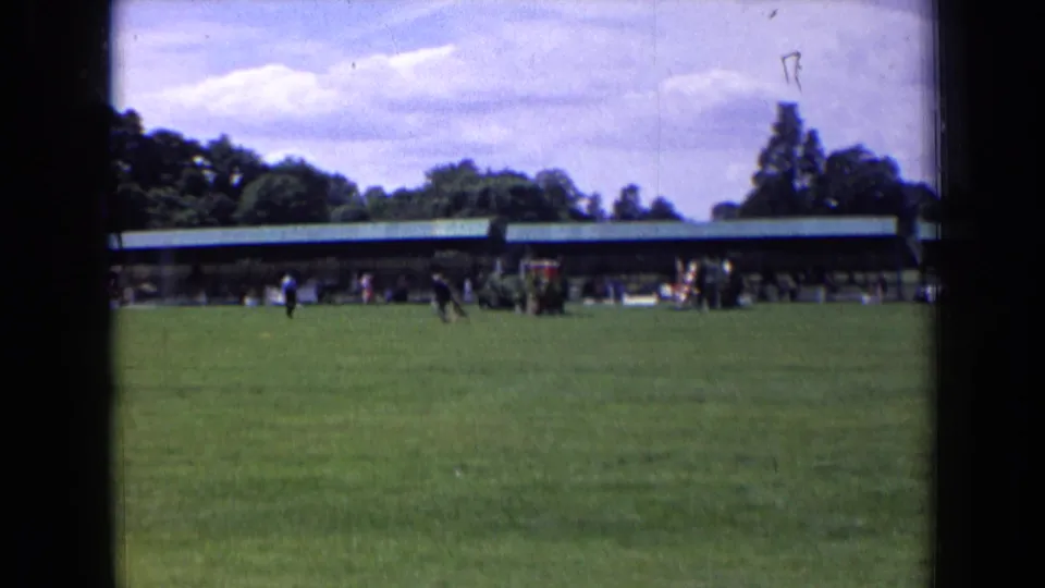 Soft 1963 England home-movie frame of a grassy field, distant people near a covered pavilion, trees, and a bright cloudy sky.