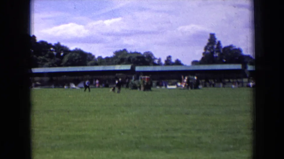 1963 England frame showing a dark-clad person and dog on a wide field, with spectators near a long green-roofed pavilion.