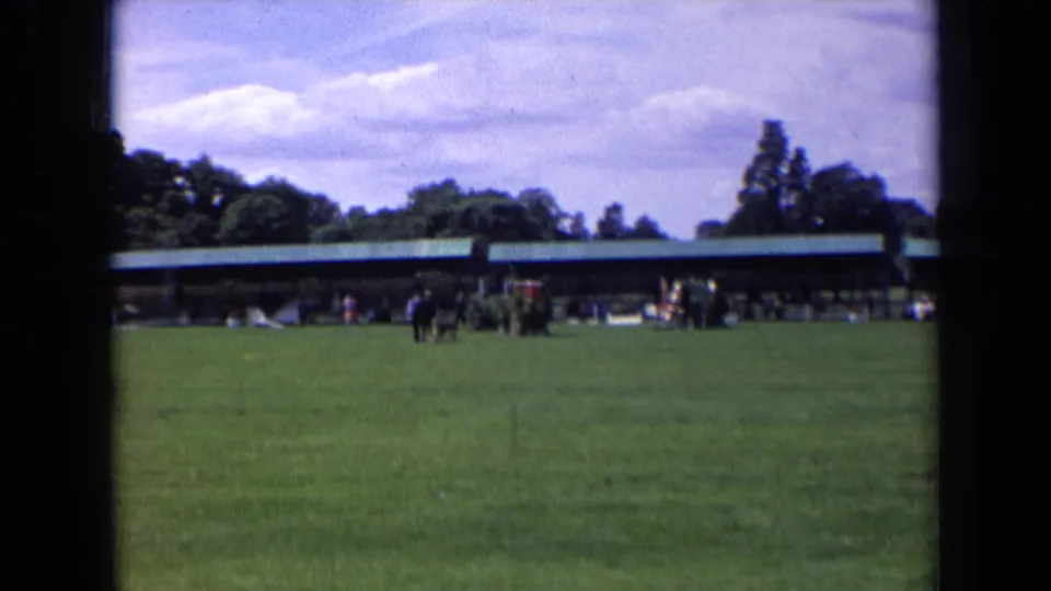 1963 England outdoor frame with people and a dog moving across a lawn, with a pavilion and trees in the background.