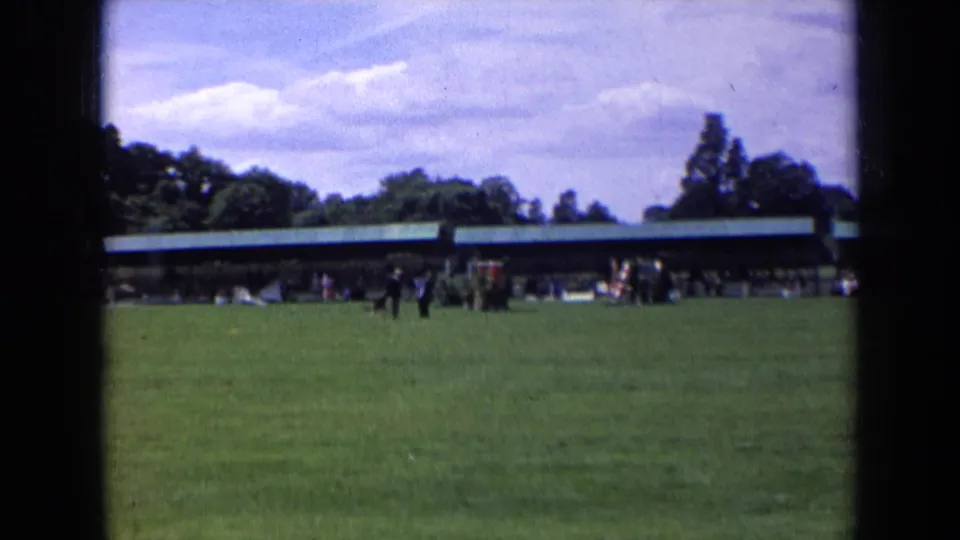 1963 England home-movie still of distant people crossing a green field near a covered pavilion, with soft 8mm color and grain.