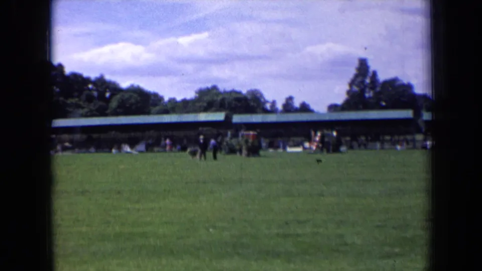 1963 England frame showing people and a dog near the center of a broad lawn, backed by a low pavilion and trees.
