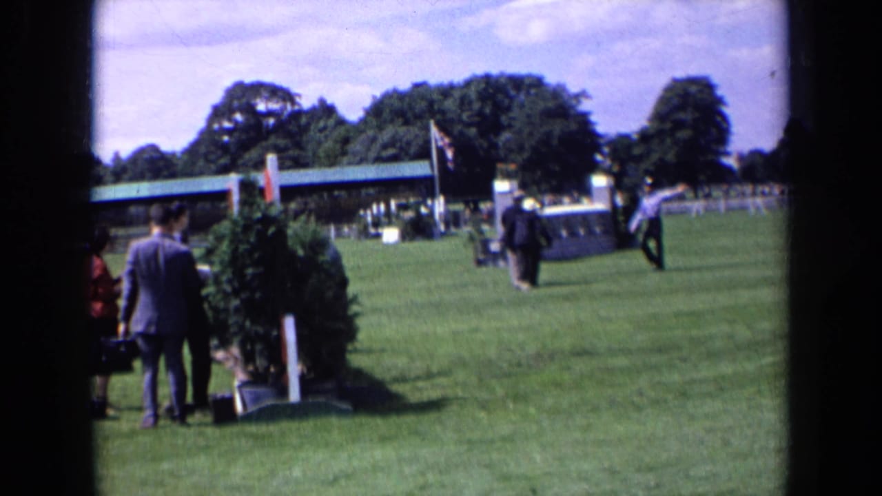 Men in suits stand on a grass field near a hedge obstacle and roofed stand.
