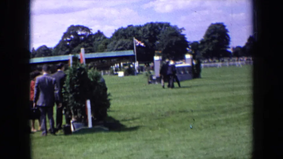 1963 England home-movie frame with men in suits moving across a showground-like grass field near flags and a low roofed structure.