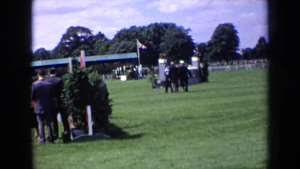1963 England 8mm frame showing suited figures in the foreground and a small group near the center of a green event field.