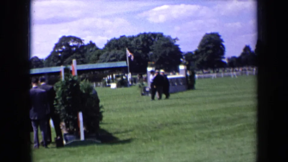 1963 England home-movie frame with suited men at the field edge and a few figures near a flagged structure in the distance.