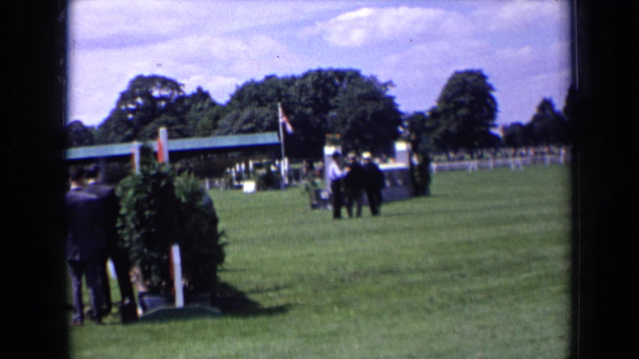 Three suited men stand or walk on a grassy field near an equestrian jump and flag.