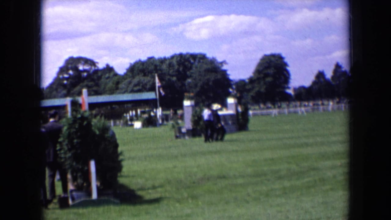 Suited men move across a sunny lawn with a flag and jump structure behind them.