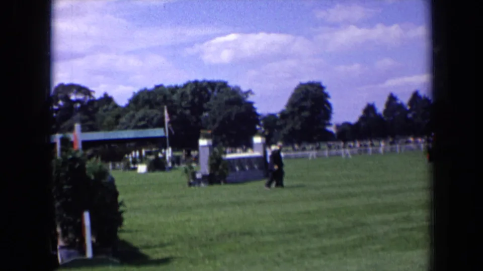 1963 England home-movie view of suited figures walking past an equestrian jump on a wide grass field under a bright sky.