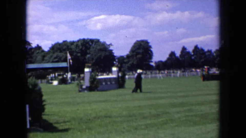 1963 England frame showing suited men crossing an event lawn with a jump obstacle, distant trees, rail fencing, and scattered clouds.