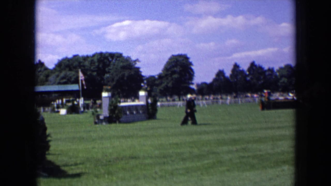 Man in a dark suit walks across a green event lawn with fencing, trees, and a flagpole behind him.